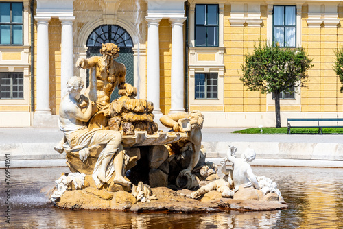 The Ehrenhofbrunnen fountain at Schonbrunn Palace stands under a clear blue sky, with water cascading over its sculptures as the palace provides an elegant backdrop. Vienna, Austria
