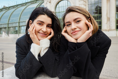 Photography cheerful lesbian couple in coats looking at camera near Palmenhaus in Vienna on