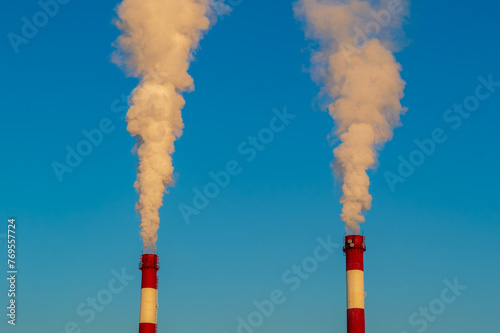 Two tall red and white smoke stacks are spewing smoke into the sky. The sky is clear and blue, with no clouds in sight. The smoke is thick and billowing, creating a sense of pollution