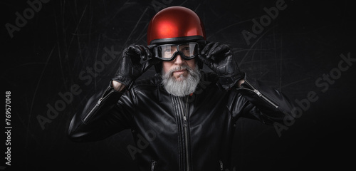 Portrait d'un motard homme avec un casque rouge et une veste en cuir et des lunettes de protection et une barbe grise vintage