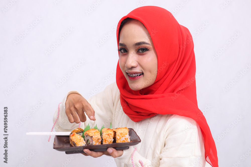 indonesia woman wearing hijab tilted to the right cheerfully looks at the camera gesturing at sushi, holding a plate containing sushi (Japanese food). Beautiful Muslim women use food and business indonesia woman wearing hijab tilted to the right cheerfully looks at the camera gesturing at sushi, holding a plate containing sushi (Japanese food). Beautiful Muslim women use food and business