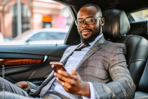 An elegantly dressed male figure utilizes a smartphone while riding in the backseat of a premium vehicle photo