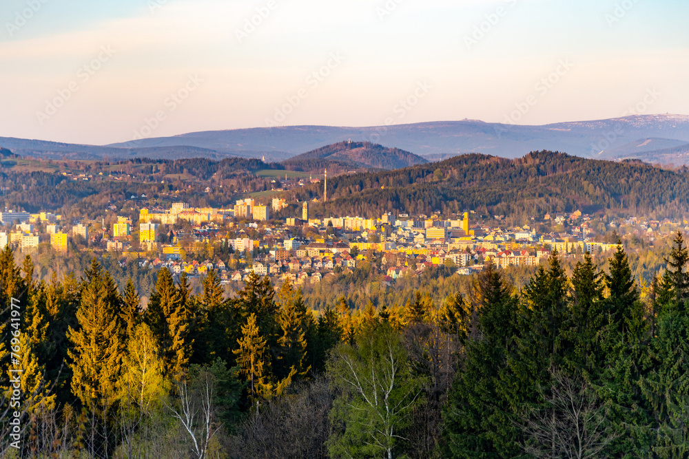The golden hour casts a warm glow over Jablonec nad Nisou, with the towering peaks of the Krkonose mountain range silhouetted in the distance, as seen through a foreground of lush greenery.