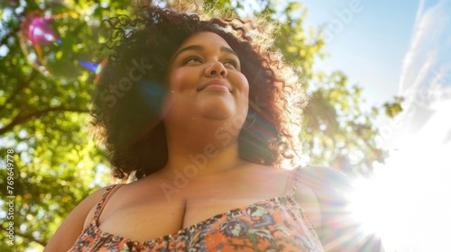 A joyful woman with curly hair wearing a floral top standing in a park with sunlight filtering through the leaves.