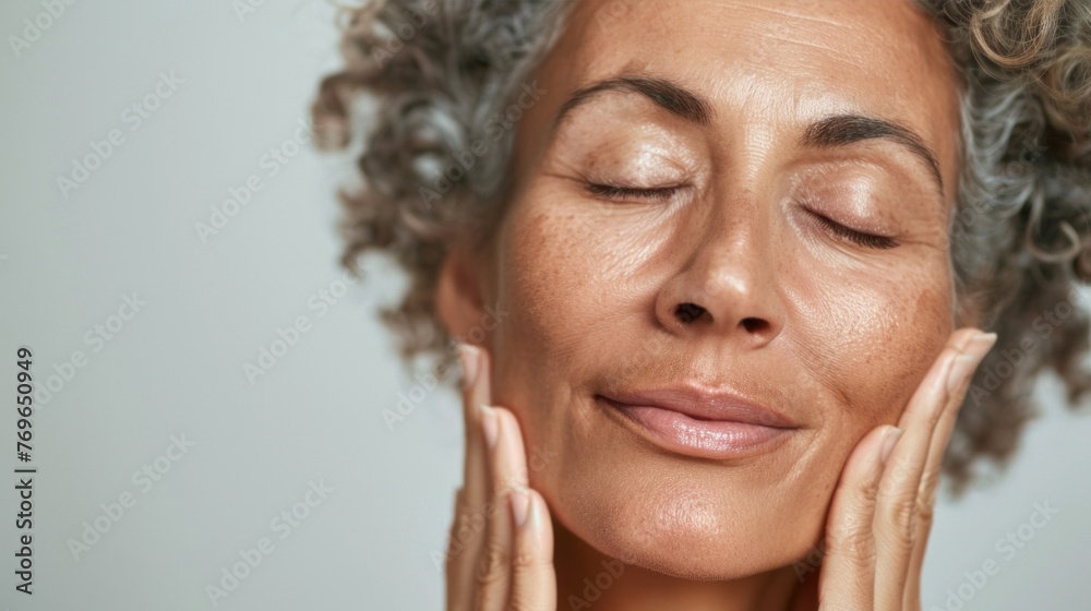 A woman with closed eyes smiling and gently massaging her face with her hands conveying a sense of relaxation and self-care. A woman with closed eyes smiling and gently massaging her face with her hands conveying a sense of relaxation and self-care.