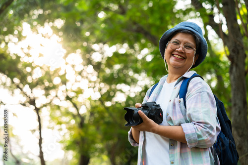 Portrait of Asian mature woman holding a camera and backpack behind her back, an Asia active senior woman enjoying nature in park. Standing on a trail in a forest outdoors. Enjoying active travel trip