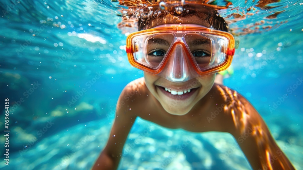 A young child underwater wearing orange goggles smiling and looking up towards the camera.