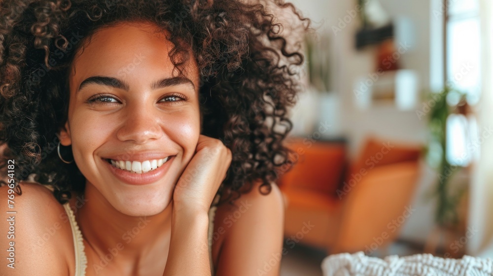 Smiling woman with curly hair resting chin on hand in warmly lit room with orange furniture and plants. Smiling woman with curly hair resting chin on hand in warmly lit room with orange furniture and plants.