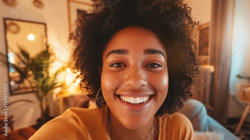 A cheerful woman with curly hair wearing a yellow top smiling brightly in a warmly lit room with plants and decorative items in the background.