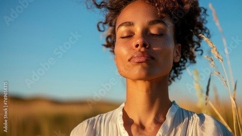 A woman with closed eyes enjoying the serenity of nature surrounded by tall grass and a clear blue sky. © iuricazac