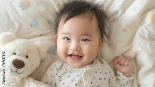 A joyful baby with a big smile lying on a patterned blanket next to a white teddy bear.