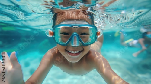 A young child with a big smile wearing blue goggles swimming underwater in a clear blue pool. © iuricazac
