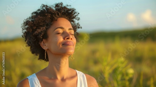 A woman with curly hair wearing a white top smiling with her eyes closed standing in a field of tall grass enjoying the moment.