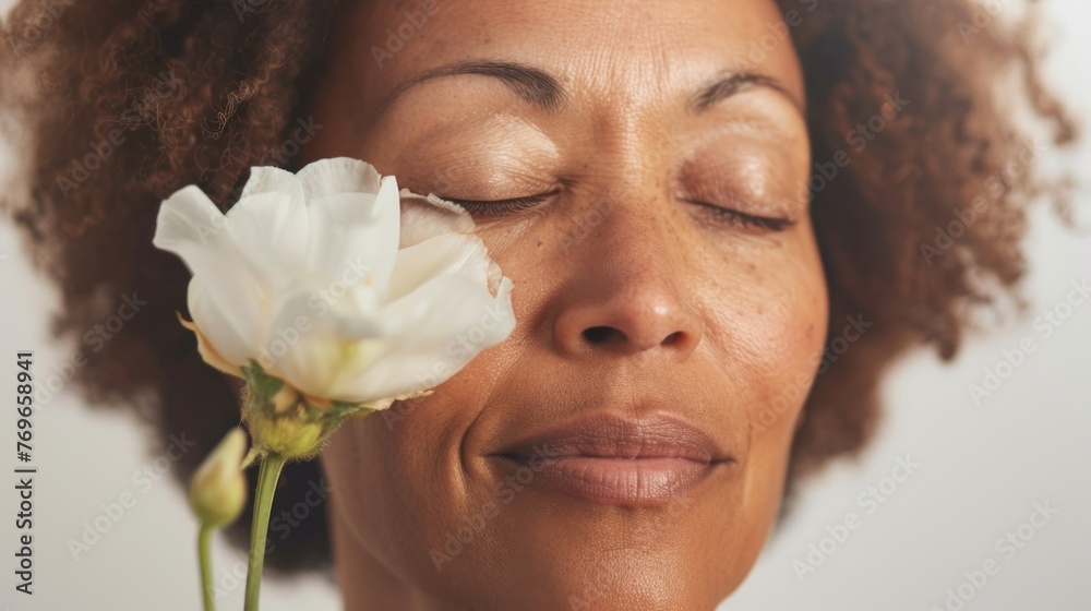 A woman with closed eyes a serene expression and curly hair smelling a white flower with a soft smile.
