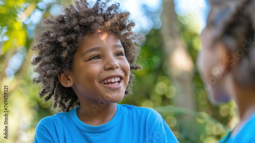 Young child with curly hair smiling at someone wearing a blue shirt outdoors with blurred greenery in the background.