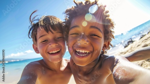 Two young boys smiling and taking a selfie on a beach with the ocean in the background.