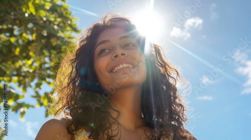 A young woman with curly hair smiling at the sun with a blurred background of a tree and blue sky.