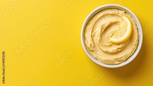 A bowl of creamy hummus topped with a lemon slice on vibrant yellow background.