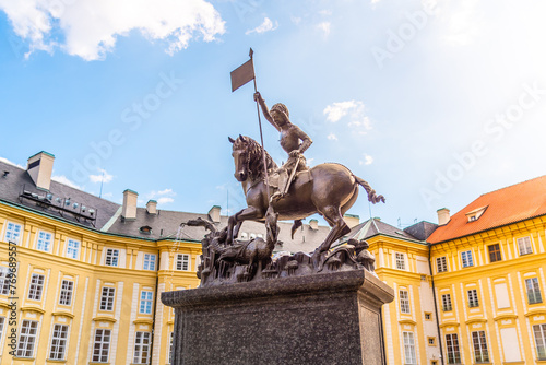 The bronze statue of Saint George triumphantly slays a dragon, set against the vibrant backdrop of Prague Castle under a blue sky. Prague, Czechia