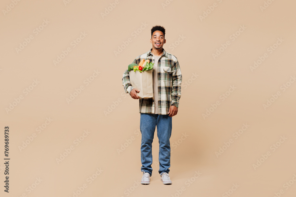 Full body smiling happy fun young man wear grey shirt hold paper bag for takeaway mock up with food products look camera isolated on plain beige background. Delivery service from shop or restaurant. Full body smiling happy fun young man wear grey shirt hold paper bag for takeaway mock up with food products look camera isolated on plain beige background. Delivery service from shop or restaurant.