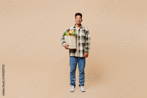Full body smiling happy fun young man wear grey shirt hold paper bag for takeaway mock up with food products look camera isolated on plain beige background. Delivery service from shop or restaurant.