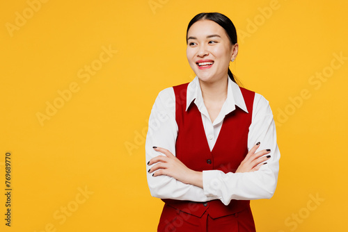 Young corporate lawyer employee business woman of Asian ethnicity wearing formal red vest shirt work at office hold hands crossed folded look aside isolated on plain yellow background. Career concept.
