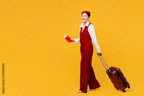 Full body young stewardess flight attendant woman of Asian ethnicity wear red vest shirt hat hold bag passport ticket travel abroad isolated on plain yellow background studio. Air flight trip concept.