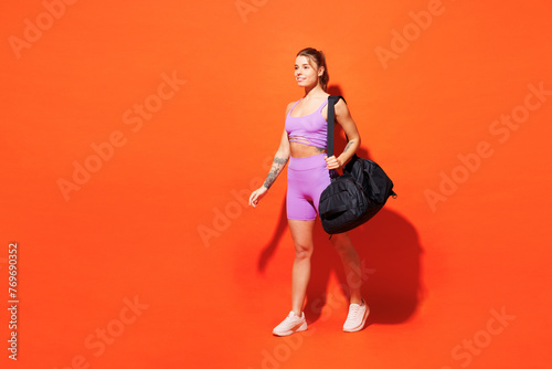 Full body sideways young smiling happy fitness trainer instructor woman sportsman wears top shorts purple clothes bag gym go to gym isolated on plain orange background. Workout sport fit abs concept.