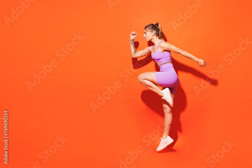 Full body side view young fitness trainer woman sportsman wear top shorts purple clothes train in home gym raise up leg going to run isolated on plain orange background. Workout sport fit abs concept. photo
