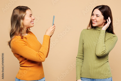 Side view young friends two women they wear orange green shirt casual clothes together doing selfie shot on mobile cell phone of buddy post photo on social network isolated on plain beige background.