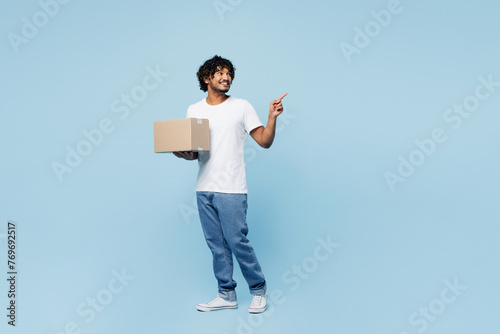 Full body young fun happy Indian man he wears white t-shirt casual clothes hold cardboard box point finger aside on area isolated on plain pastel light blue cyan background studio. Lifestyle concept.