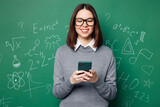Young smart teacher woman in grey casual shirt glasses hold use mobile cell phone search information isolated on plain green wall chalk blackboard background Education in high school college concept