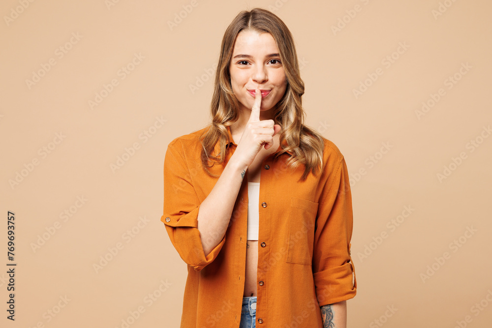 Young secret Caucasian woman she wear orange shirt casual clothes say hush be quiet with finger on lips shhh gesture isolated on plain pastel light beige background studio portrait. Lifestyle concept. - obrazy, fototapety, plakaty 