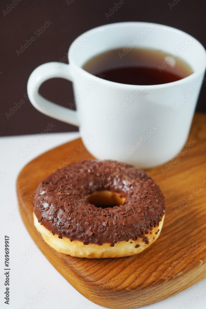 Donut with chocolate icing next to white cup of tea on wooden board on white and brown background