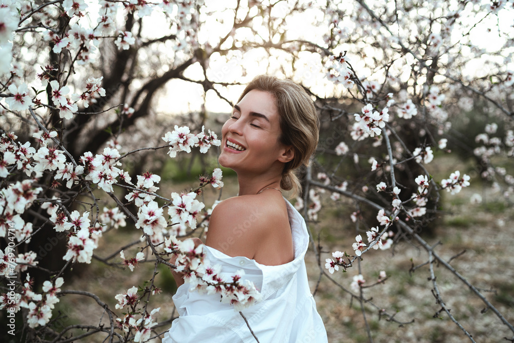 Beautiful young woman wearing a white shirt, elegantly posing near the blossoming almond trees Beautiful young woman wearing a white shirt, elegantly posing near the blossoming almond trees