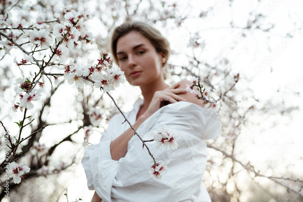 Beautiful young woman wearing a white shirt, elegantly posing near the blossoming almond trees Beautiful young woman wearing a white shirt, elegantly posing near the blossoming almond trees