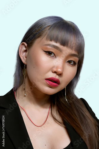 Close-up portrait of a woman with striking makeup, featuring a bold lip color and detailed eye makeup, complemented by a black outfit and statement earrings against a light blue background.