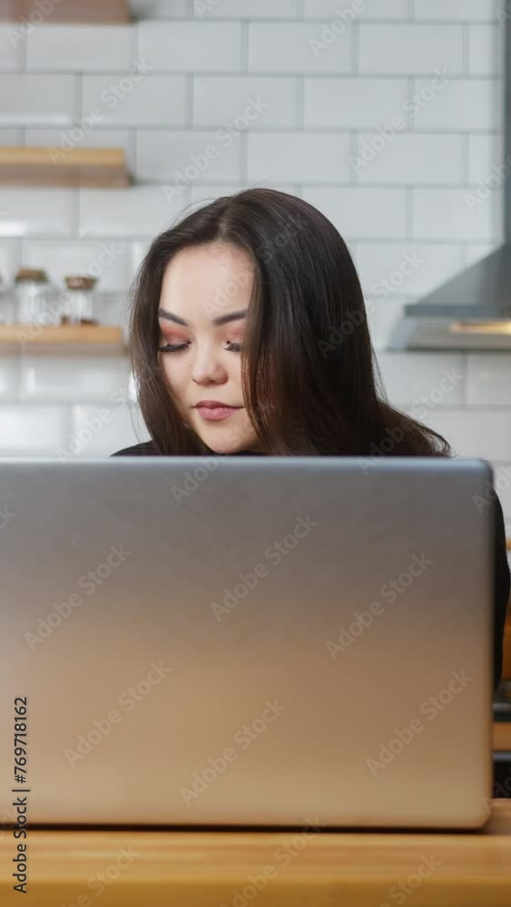 Vertical video. Asian woman work at home in the kitchen working with his laptop, taking notes, doing research. Busy freelancer working on modern tech notebook device	
