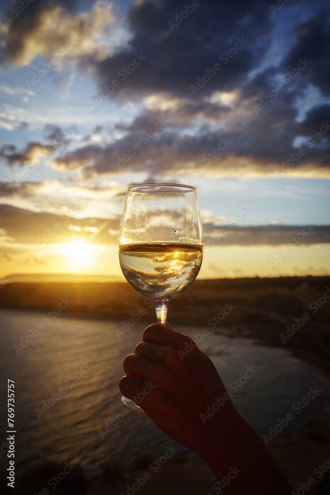 Female hand with glass of white wine against a breathtaking seaside sunset