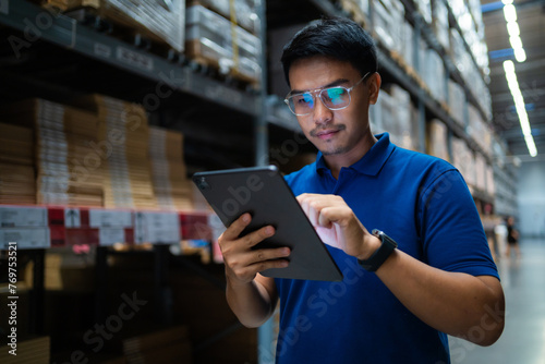 A man wearing glasses is looking at a tablet in a warehouse. He is focused on the screen and he is working on something