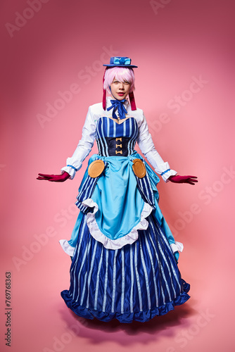 fashionable chic female cosplayer in blue hat and vivid dress looking at camera on pink backdrop
