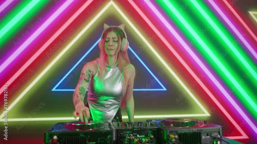Attractive female on digital wall colorful visual graphic background. Young woman dj dancing, plays music on turntables mixer at night club, looks at camera.