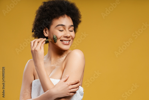 Young African American woman in white towel after shower doing facial massage, using massage roller
