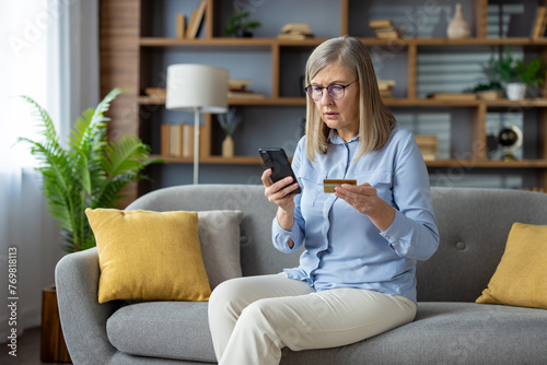 Foto Anxious mature woman sitting on sofa, holding credit card and phone, looking at screen with concern, suspecting a fraudulent transaction