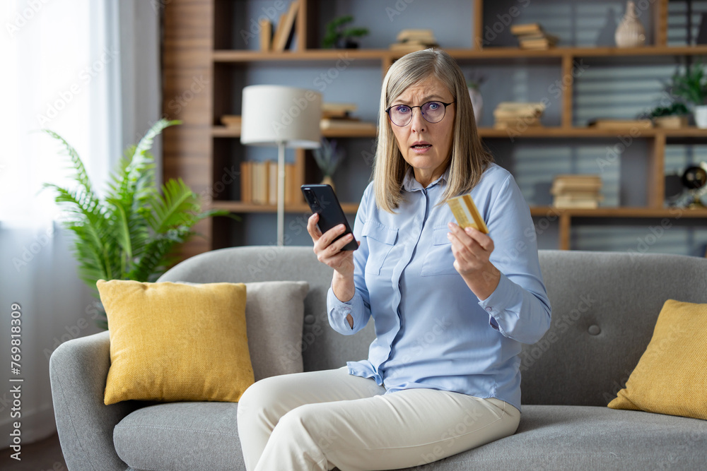 A mature adult woman examines her credit card while holding a smartphone, sitting comfortably on her couch at home, possibly suspicious of fraud. A mature adult woman examines her credit card while holding a smartphone, sitting comfortably on her couch at home, possibly suspicious of fraud.