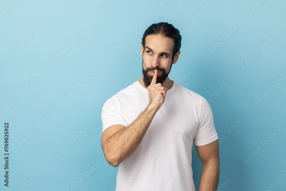 Shh, it's big secret. Portrait of man with beard wearing white T-shirt smiling, showing gesture secret sign with finger near his lips. Indoor studio shot isolated on blue background. Shh, it's big secret. Portrait of man with beard wearing white T-shirt smiling, showing gesture secret sign with finger near his lips. Indoor studio shot isolated on blue background.
