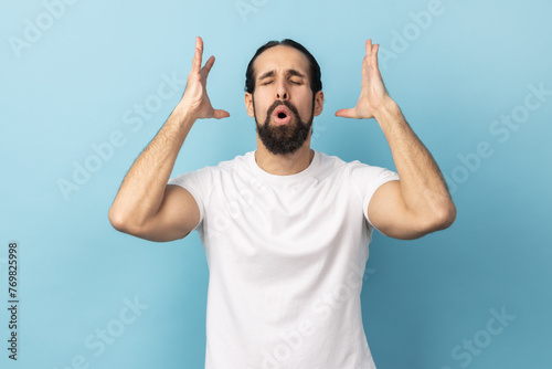 Portrait of man with beard wearing white T-shirt touching his head and showing explosion, looking worried and shocked, deadline, professional burnout. Indoor studio shot isolated on blue background. photo