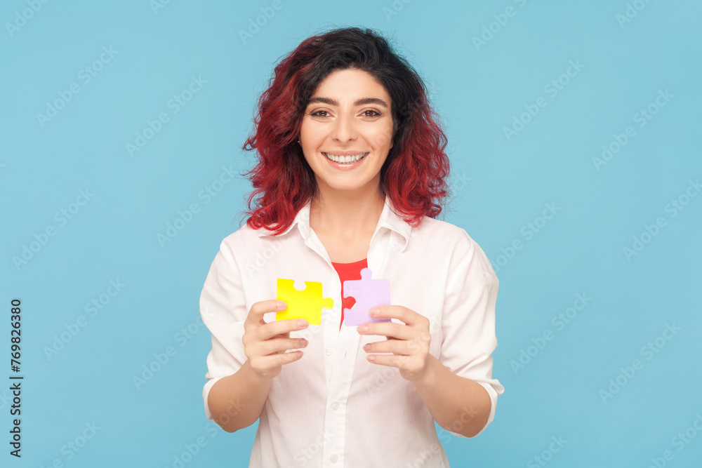 Portrait of smiling beautiful friendly woman with fancy red hair holding piece of puzzles, solving tasks, answering questions, wearing white shirt. Indoor studio shot isolated on blue background. Portrait of smiling beautiful friendly woman with fancy red hair holding piece of puzzles, solving tasks, answering questions, wearing white shirt. Indoor studio shot isolated on blue background.