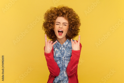 Portrait of excited woman with Afro hairstyle depicting heavy metal rock sign, horns up gesture, screaming with widely open mouth. Indoor studio shot isolated on yellow background.