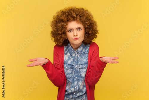 What do you want? Portrait of woman with Afro hairstyle raising arms in questioning gesture, saying I don't understand problem, looking indignant. Indoor studio shot isolated on yellow background.